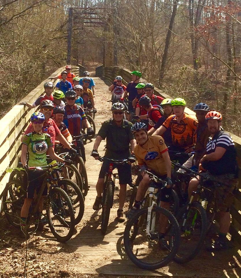 A group of mountain bikers gathered on a wooden bridge in a wooded area, smiling and posing for the camera. The scene features a diverse group of people wearing helmets and colorful cycling jerseys, with bicycles parked alongside them. The trees are bare, indicating early spring, and there is a clear sky in the background.