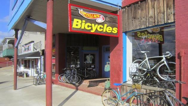 An exterior view of a bicycle shop named "Cycle Path Bicycles." The shop features a bright red sign displaying the name, with a variety of bicycles parked outside. The building has a rustic wooden facade and is situated in a small town setting with blue skies in the background.