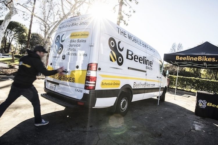 A white van with yellow and black branding for Beeline Bikes parked in a sunny area. A person in a black jacket is approaching the van, which is labeled "THE MOBILE BIKE SHOP." A tent with the Beeline Bikes logo and website is set up nearby, along with a black table displaying promotional materials. Trees and clear blue sky are visible in the background.