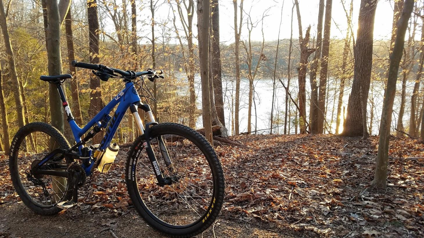 Kona Process 134: A blue mountain bike is positioned on a leaf-covered trail in a wooded area. The background features tall trees and a glimmering river reflecting sunlight, creating a peaceful outdoor scene during early evening.