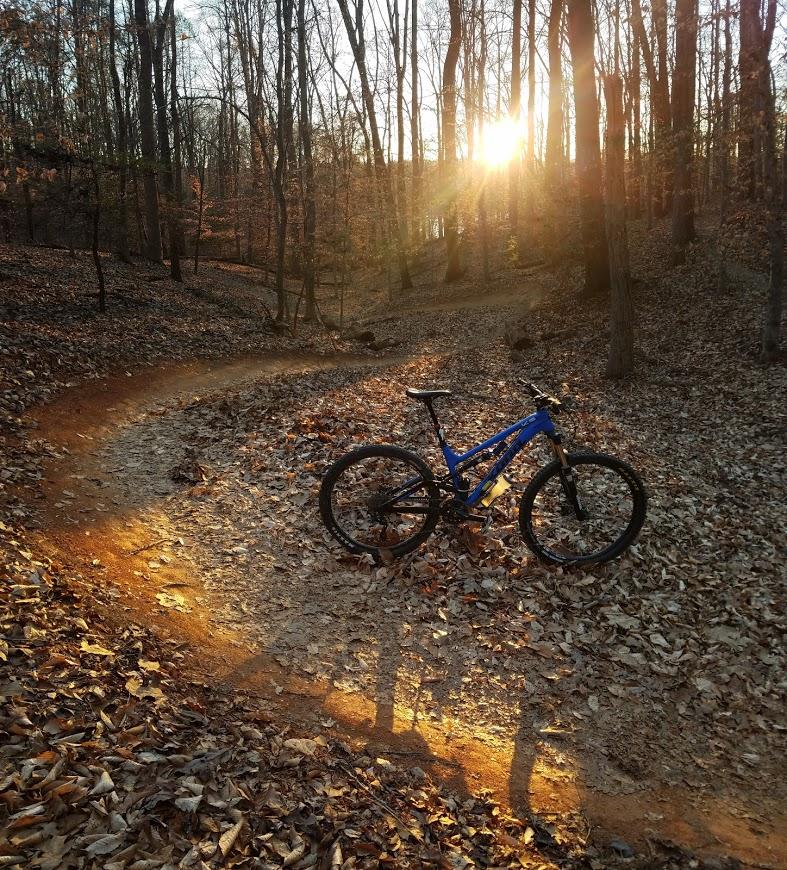 A blue mountain bike is resting next to a winding dirt path in a forest during sunset. The ground is covered in fallen leaves, and the warm glow of the setting sun filters through the trees, casting long shadows. Fountainhead Regional Park mountain bike trail.
