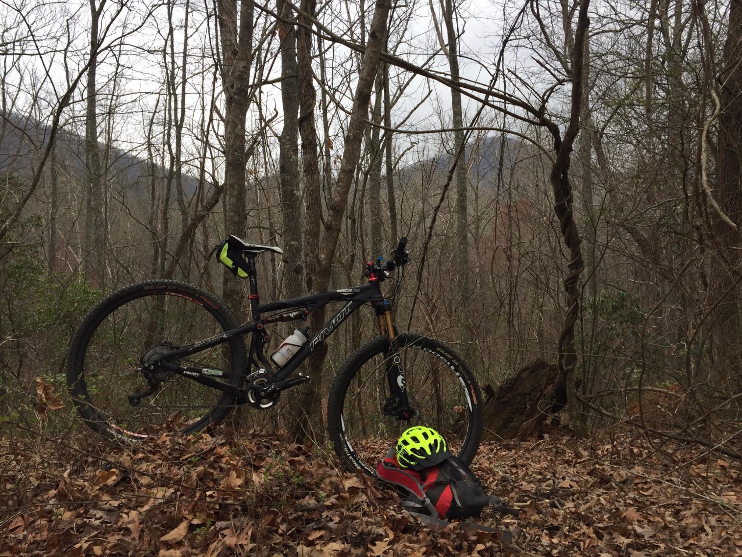 Pivot Mach 429: A mountain bike with a water bottle attached is parked beside a red and black backpack on the ground, with a bright yellow helmet resting on top. The scene is set in a forested area during cloudy weather, surrounded by bare trees and fallen leaves. In the background, faint mountain silhouettes are visible.