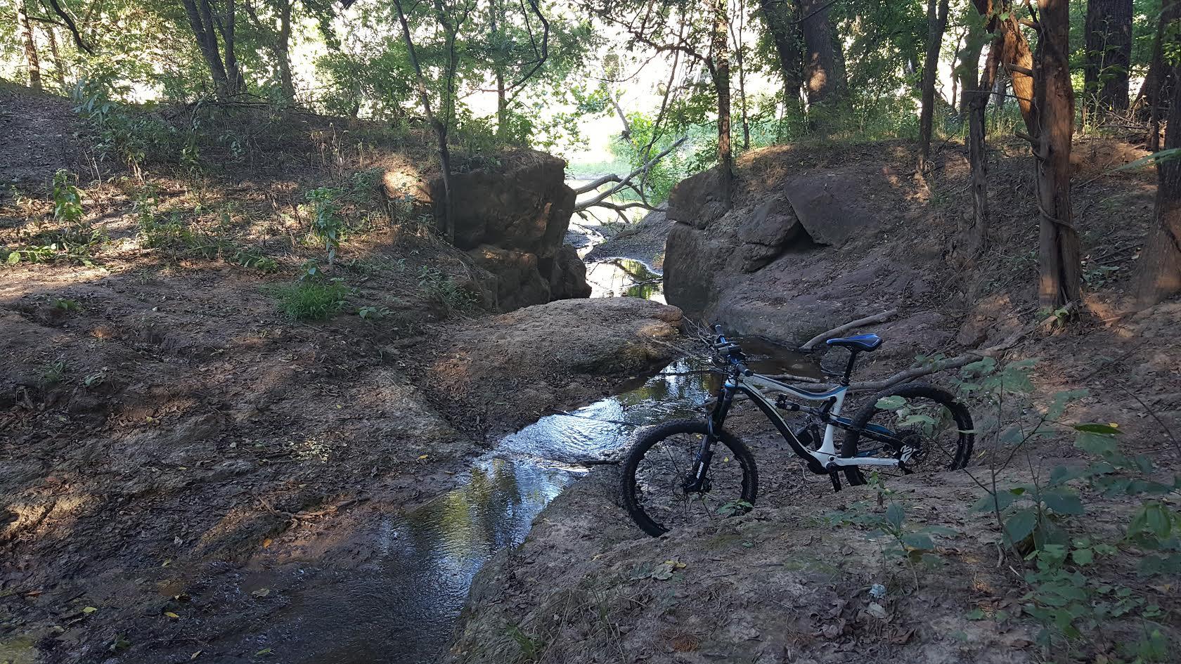 A mountain bike parked near a shallow stream surrounded by rocky terrain and trees in a wooded area. Sunlight filters through the foliage, illuminating the natural landscape. Devil