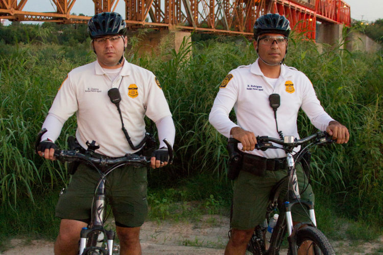 Two border patrol agents stand beside their bicycles in a grassy area near a bridge. They are wearing uniforms with badges and helmets, with communication devices attached to their shirts. The background features tall grass and the silhouette of an orange bridge.