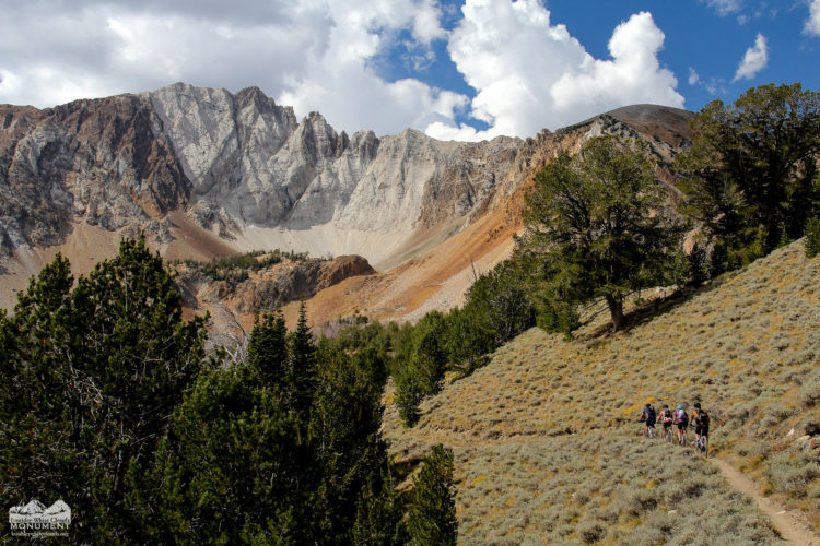 A scenic view of a hiking trail winding through a grassy area with shrubs, leading towards a mountainous backdrop featuring rugged, rocky cliffs and a clear blue sky with scattered clouds. Multiple hikers are visible on the trail, surrounded by evergreen trees.