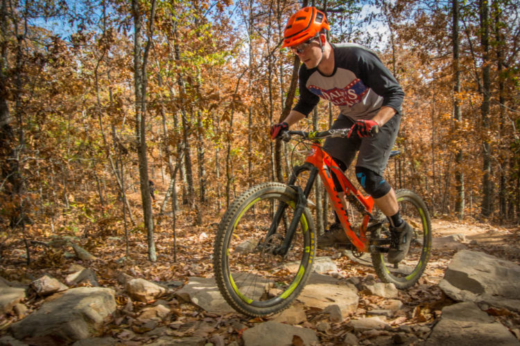 A mountain biker navigating a rocky trail in a wooded area during autumn. The scene features colorful fall foliage and a clear blue sky, while the rider, wearing a helmet and protective gear, demonstrates skill and focus as he rides over the uneven terrain.