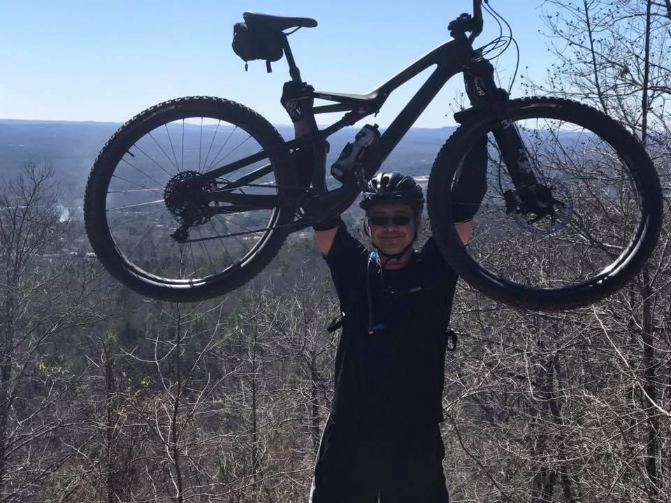 A person in a black helmet and riding gear is joyfully holding a mountain bike above their head while standing on a hilltop, overlooking a valley with trees in the background and a clear blue sky. Coldwater Mountain mountain bike trail.