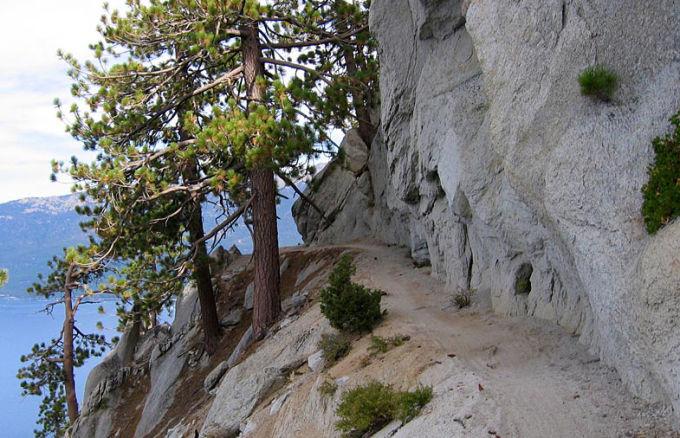 A narrow trail winding along a rocky cliffside, surrounded by pine trees, with a view of a blue lake in the background. The landscape features rugged grey rock formations and patches of green vegetation. Flume Trail mountain bike trail.