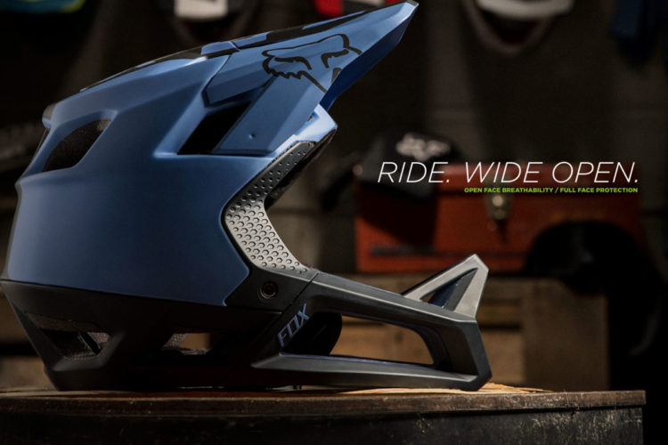 A blue full face helmet with an open face design, featuring ventilation holes and the Fox Racing logo, positioned on a wooden surface. The text "RIDE. WIDE OPEN." is displayed prominently next to the helmet, highlighting its breathability and protective features. A blurred background includes various biking accessories and gear.