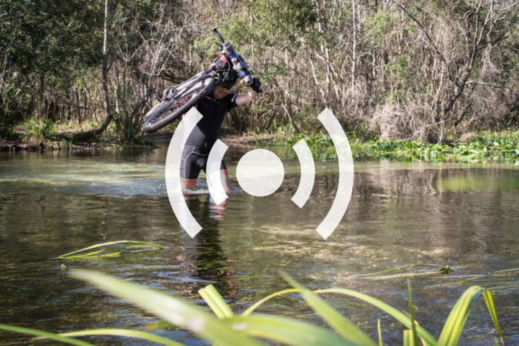 A person wading through shallow water in a natural setting, carrying a bicycle above their head. The surrounding landscape features trees and greenery, creating an outdoor adventure scene.