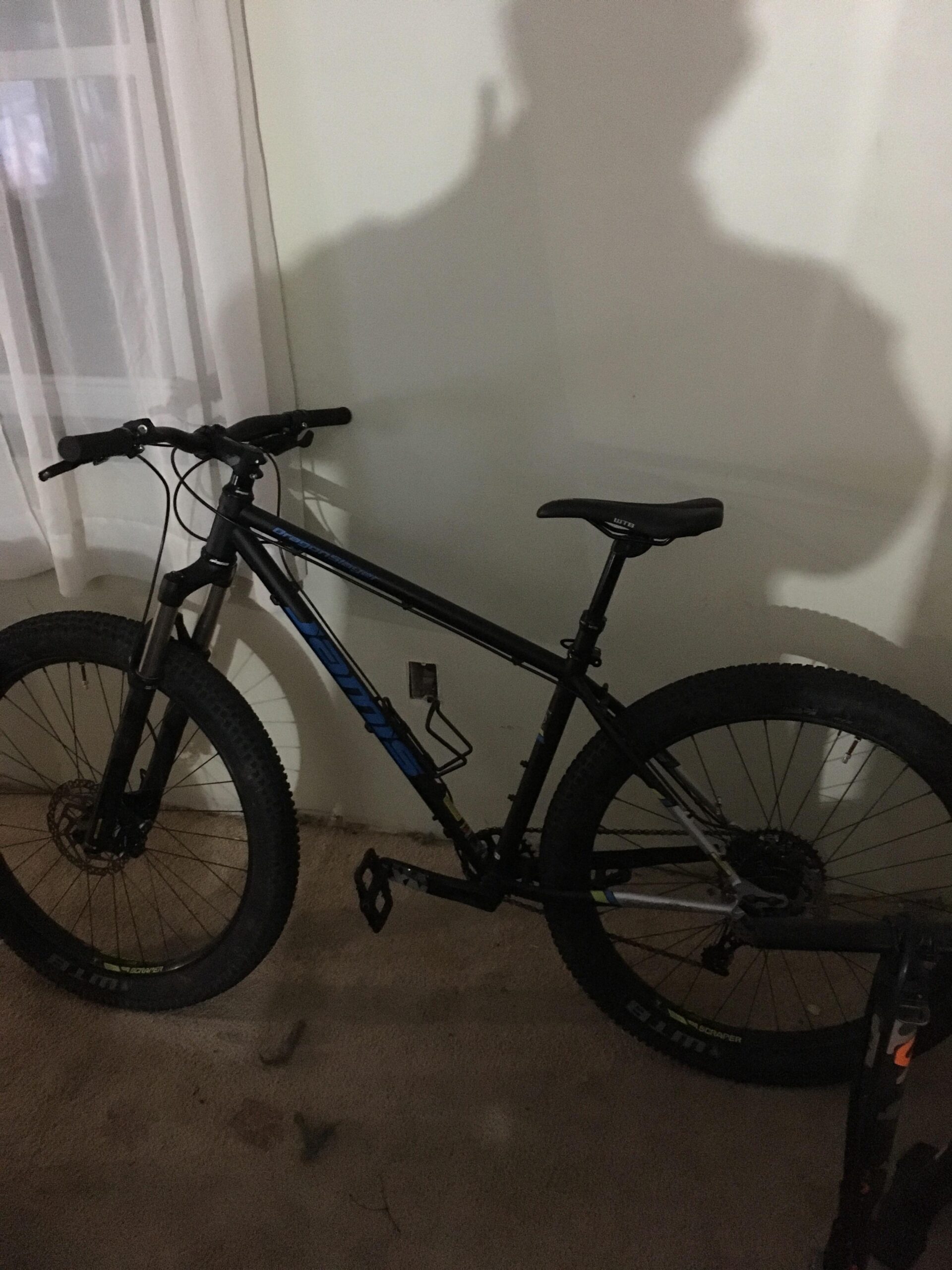 Jamis Dragonslayer 27.5: A black mountain bike with blue accents is leaning against a wall in a dimly lit room, casting a shadow. The bike features thick tires and a simple design, with the handlebars and seat clearly visible. Light filters through a sheer curtain, creating a soft atmosphere in the background.