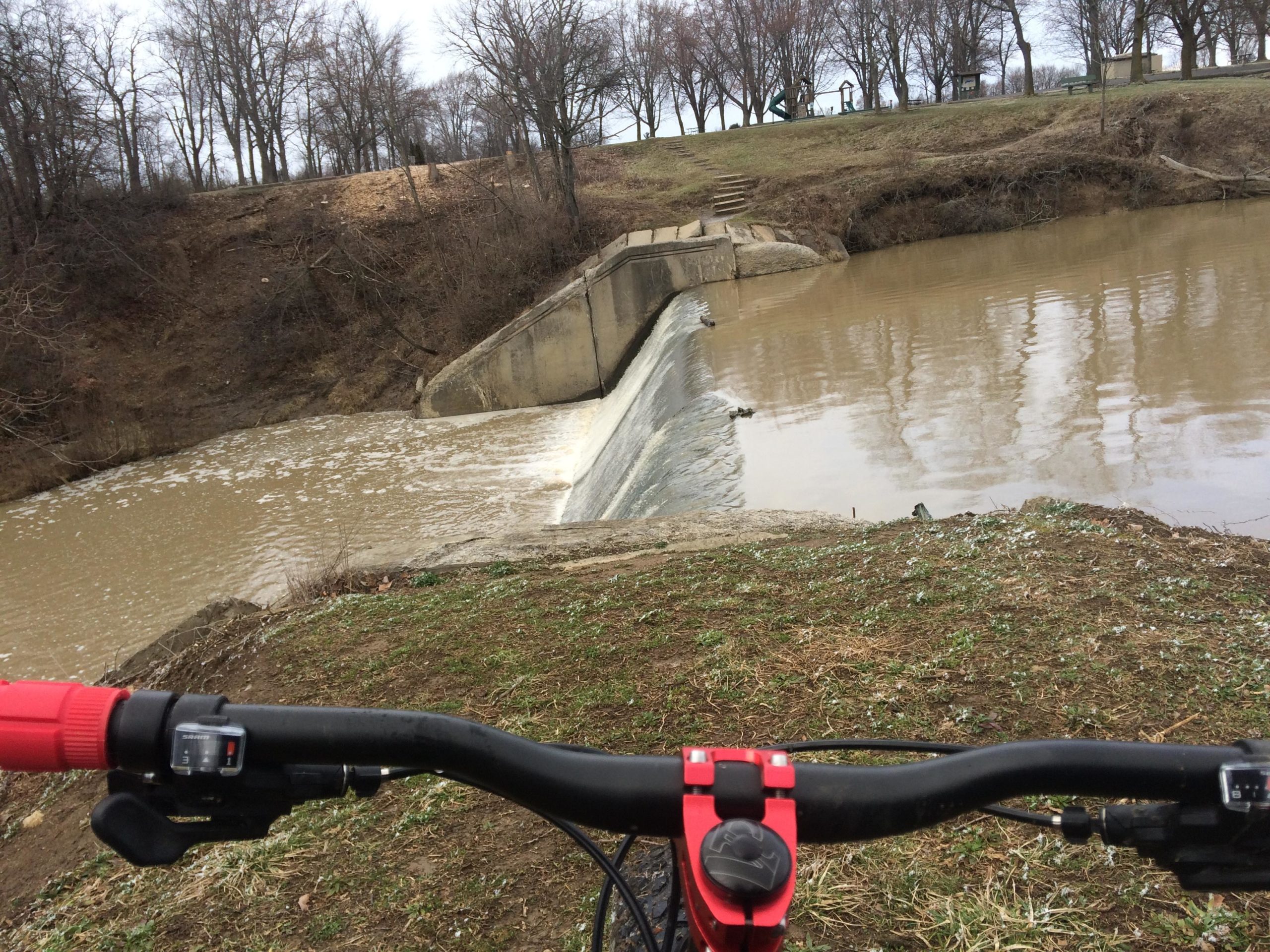 A view from the handlebars of a bicycle overlooking a brown, murky body of water with a concrete spillway on the right. In the background, trees without leaves are visible along a hill, and there are steps leading up from the water's edge. The scene is cloudy and appears to be early spring or late winter. Van  Buren mountain bike trail.