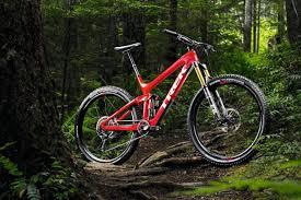 Trek Slash 9.9: A vibrant red mountain bike positioned on a dirt trail in a lush, green forest. The bike features wide tires and a robust frame, surrounded by tall trees and foliage, highlighting a scenic outdoor environment.