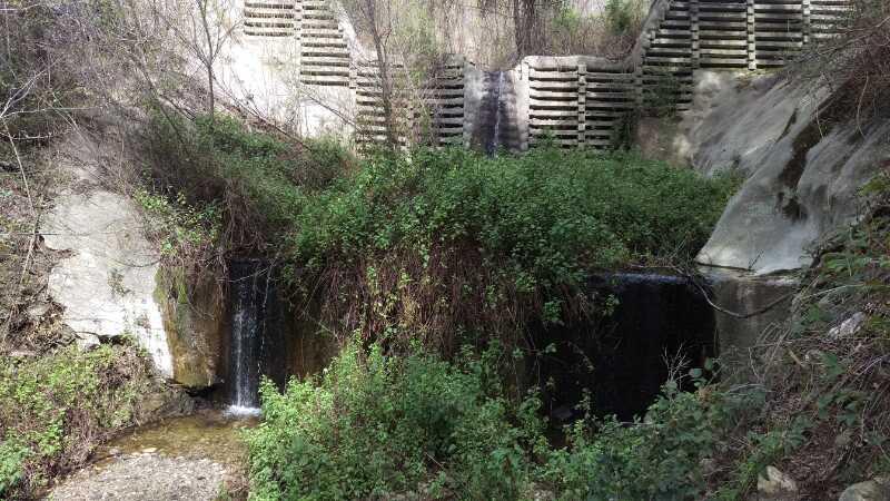 A small waterfall flowing through lush greenery, with concrete walls and wooden structures in the background. Bare trees and dense plants surround the water, creating a natural yet urban landscape. El Prieto mountain bike trail.
