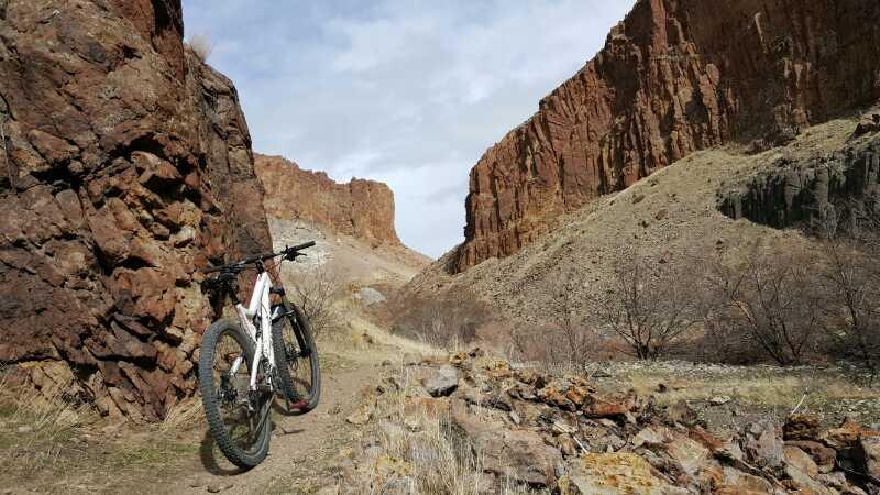 A mountain bike leaning against a rocky path surrounded by tall cliffs and barren terrain under a cloudy sky. Wilson Creek Area mountain bike trail.