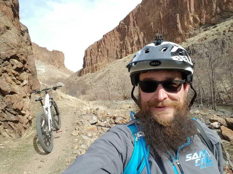A person with a beard wearing a helmet and sunglasses smiles for a selfie on a mountain biking trail, with rocky cliffs and a white mountain bike in the background. The setting appears to be a dry, outdoor landscape. Wilson Creek Area mountain bike trail.