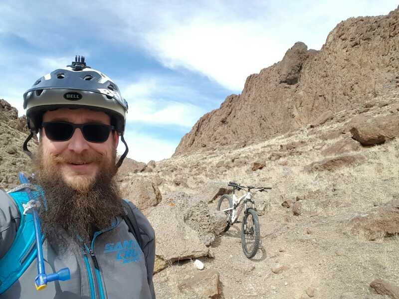 A man wearing a helmet and sunglasses smiles for a selfie while standing on a rocky trail, with a mountain bike parked behind him. The landscape features rugged terrain and scattered rocks under a blue sky with clouds. Wilson Creek Area mountain bike trail.