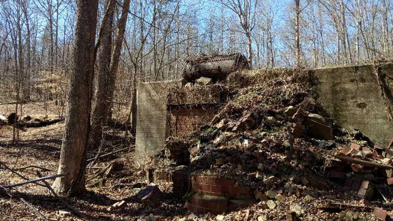 Alt tag: "Ruins of a collapsed structure covered in foliage and broken bricks, surrounded by bare trees in a forest setting." Chicopee Woods mountain bike trail.