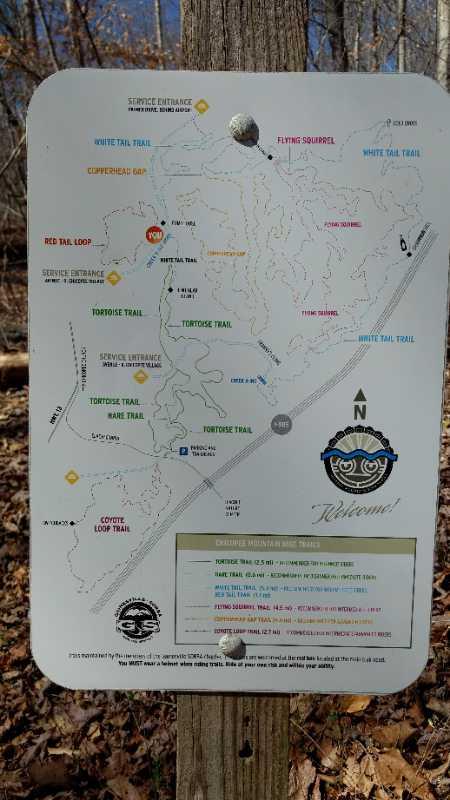 Map of a nature trail system showing various hiking routes, including Red Tail Loop, Tortoise Trail, and White Tail Trail. The map includes symbols for trails, points of interest, and an overview of trail lengths and difficulty levels, with a compass rose indicating direction. The sign is mounted on a wooden post, surrounded by trees and forest floor. Chicopee Woods mountain bike trail.