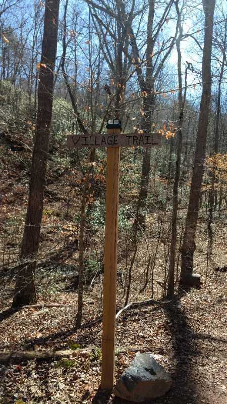 A wooden trail sign marking the "Village Trail" stands in a forested area, surrounded by bare trees and fallen leaves, with sunlight filtering through the branches. Chicopee Woods mountain bike trail.