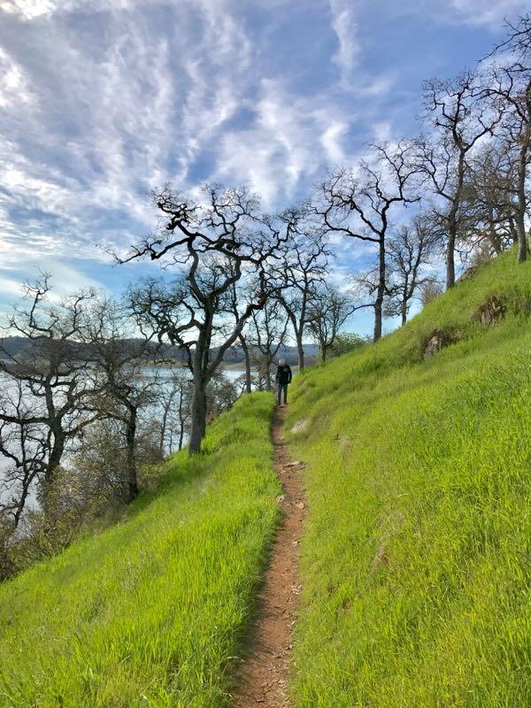 A person walking along a narrow dirt path bordered by lush green grass and trees, with a serene lake visible in the background under a partly cloudy sky. Glory Hole mountain bike trail.