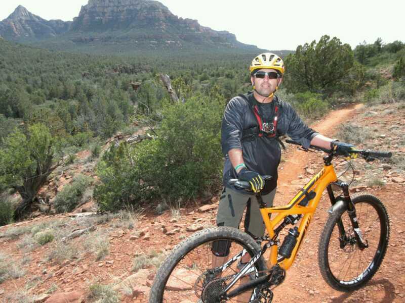 A mountain biker wearing a yellow helmet and sunglasses stands next to an orange bike on a dirt trail surrounded by lush greenery and rocky mountains in the background. The rider is dressed in a long-sleeve shirt and padded shorts, ready for an outdoor adventure. Chuck Wagon mountain bike trail.