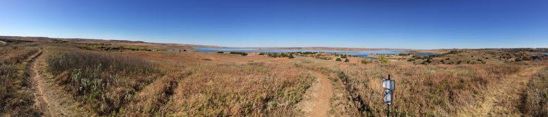 A panoramic view of a scenic landscape featuring a wide, open field with tall grass and wildflowers, alongside a winding dirt path. In the distance, a calm lake reflects the clear blue sky, while rolling hills frame the horizon. A wooden sign stands next to the path, indicating directional information. The scene conveys a peaceful, natural environment ideal for outdoor exploration. Switchgrass mountain bike trail.