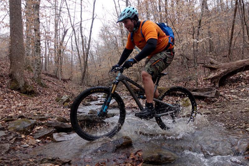 A mountain biker in an orange shirt and helmet rides through a shallow stream, splashing water as he navigates over rocks, surrounded by a forest with bare trees and fallen leaves. Upper Buffalo Headwaters Trail System mountain bike trail.