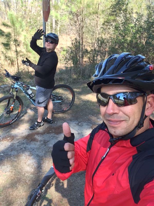 Two cyclists take a selfie while enjoying a ride on a wooded trail. One cyclist is giving a thumbs-up while the other waves, both wearing helmets and sunglasses. A stop sign is visible in the background, surrounded by trees and natural scenery. Blue Clay Bike Park mountain bike trail.