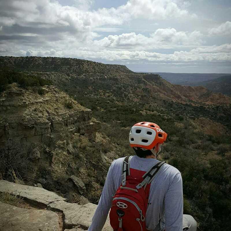 A person wearing a red and white helmet sits on a rocky ledge, looking out over a vast canyon landscape under a cloudy sky. The scene features rolling hills and a mix of vegetation, showcasing the natural beauty of the area. Palo Duro Canyon mountain bike trail.