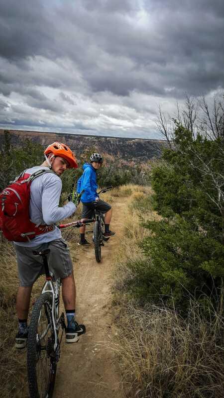 Two mountain bikers pause on a narrow dirt trail surrounded by dry grass and shrubs. One biker, wearing an orange helmet and a red backpack, looks back at the camera, while the other, dressed in a blue jacket, stands slightly ahead. The scene is set against a backdrop of cloudy skies and a canyon in the distance, suggesting an adventurous outdoor activity. Palo Duro Canyon mountain bike trail.