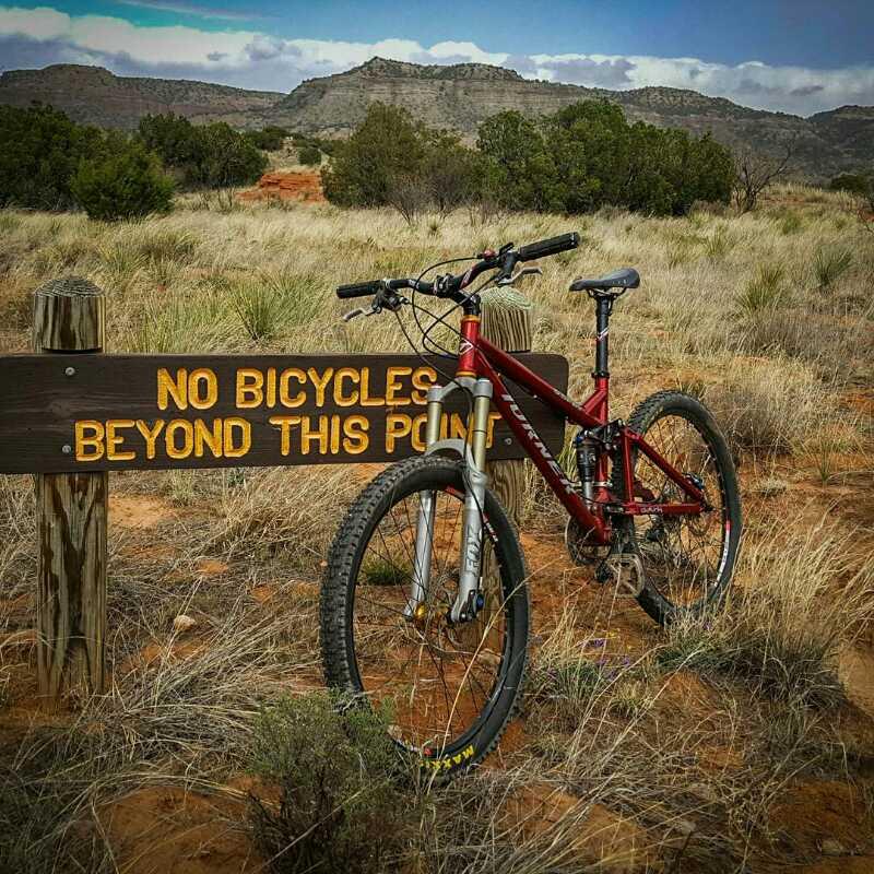 A red mountain bike rests against a wooden sign that reads "No Bicycles Beyond This Point," with a natural landscape of grass and shrubs in the background, set against rocky hills under a partly cloudy sky. Palo Duro Canyon mountain bike trail.