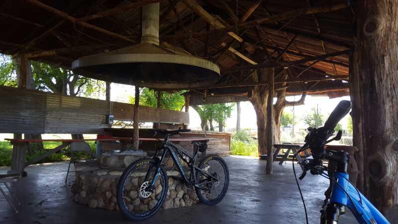 A rustic outdoor pavilion with a thatched roof, featuring wooden beams and a stone fireplace at the center. Two mountain bikes are parked in the foreground, and picnic tables are visible in the background, surrounded by greenery. The space is well-lit with natural sunlight filtering through the trees. Rocky Hill Ranch mountain bike trail.