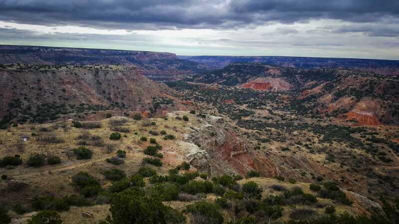 A panoramic view of rugged canyon landscape under a cloudy sky, featuring steep cliffs, scattered shrubs, and varying shades of earthy tones, highlighting the natural beauty of the terrain. Palo Duro Canyon mountain bike trail.