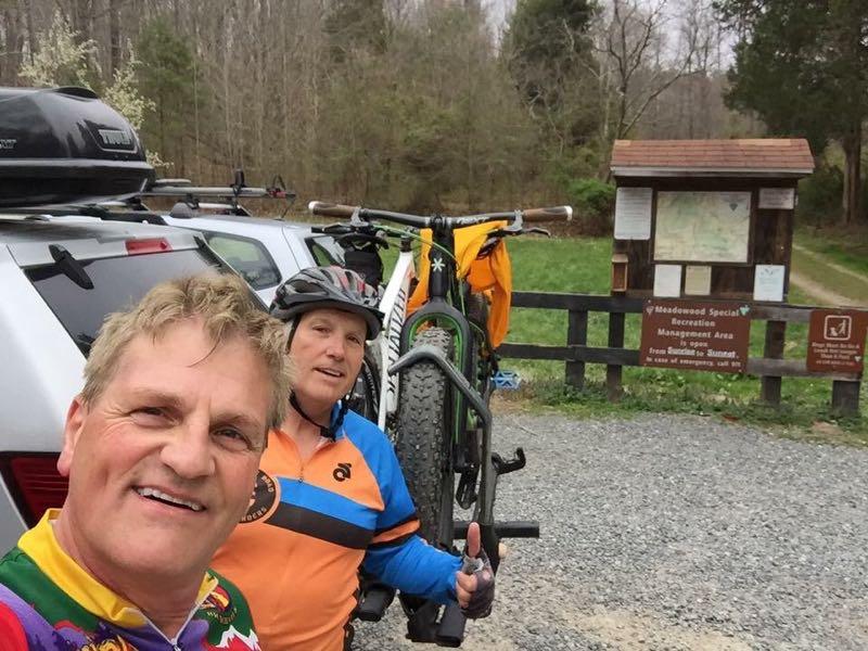 Two men pose for a selfie in front of a vehicle parked near a trailhead. They are wearing colorful cycling jerseys and helmets, with bicycles mounted on the back of the car. In the background, there is a wooden sign indicating the trail area, surrounded by trees and a gravel path. Meadowood mountain bike trail.