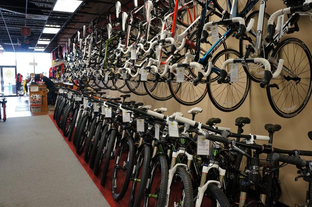 A well-lit bike shop interior featuring a wide selection of bicycles. The wall displays numerous road bikes hanging on hooks, while a row of mountain bikes lines the floor, each equipped with price tags. A salesperson can be seen at the counter in the background, assisting a customer.