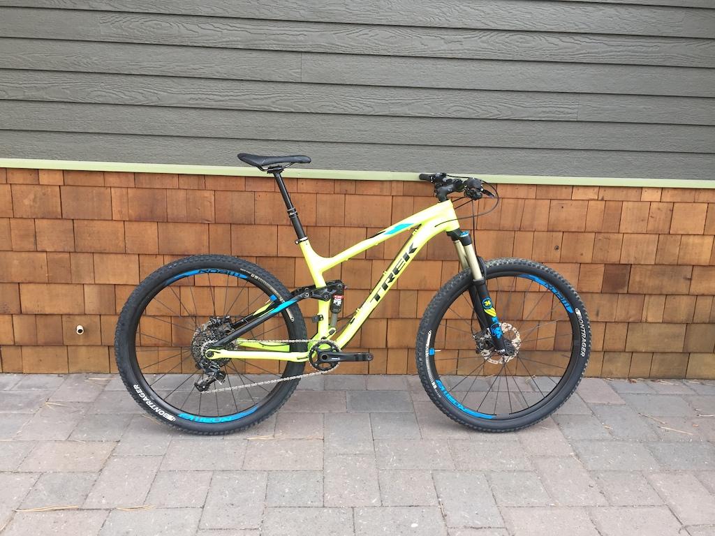 Trek Fuel EX 9 29: A colorful yellow and black mountain bike with blue accents, positioned against a wooden wall. The bike features a front suspension fork and disc brakes, highlighting its design for off-road cycling. The background consists of a textured wooden surface, adding contrast to the bike.
