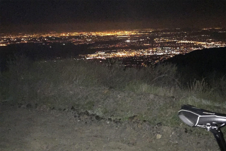 A nighttime view from a hillside overlooking a city, illuminated by countless lights. In the foreground, there's a portion of a dirt path with grass and shrubs, and the seat of a bicycle is visible to the right, adding context to the scene.