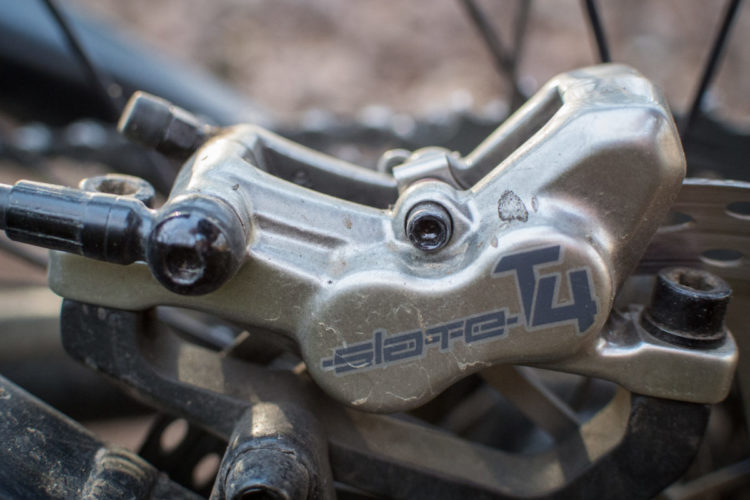 Close-up of a bike brake caliper, showcasing its metallic finish and components. The image features the branding "SLATE T4," with visible details of the brake lines and attachment points, set against a blurred background of bicycle parts.