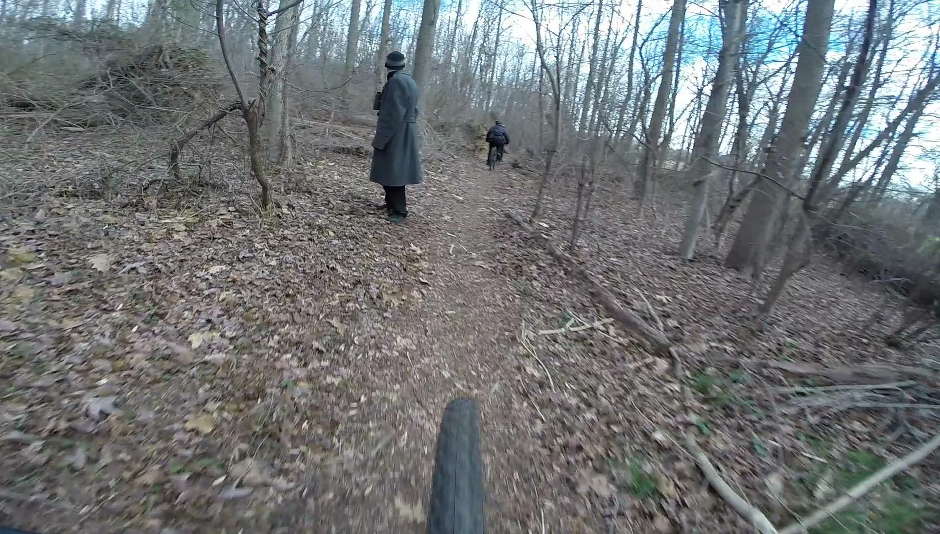 A view along a narrow trail in a wooded area during winter, with leafless trees in the background. Two figures are present: one standing in a long coat and hat, and another riding a bicycle further down the path. The ground is covered with fallen leaves and twigs, creating a natural and rustic atmosphere. Richmond Avenue and Forest Hill road mountain bike trail.