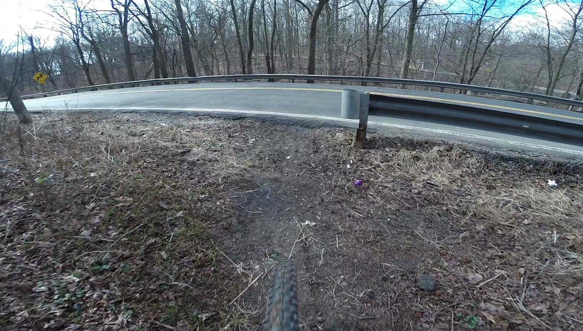 A close-up view of a bike tire on a dirt path beside a winding road, surrounded by bare trees in early spring. The image captures the texture of the ground and a portion of the road with a curve, indicating a natural outdoor setting for cycling or mountain biking. Richmond Avenue and Forest Hill road mountain bike trail.