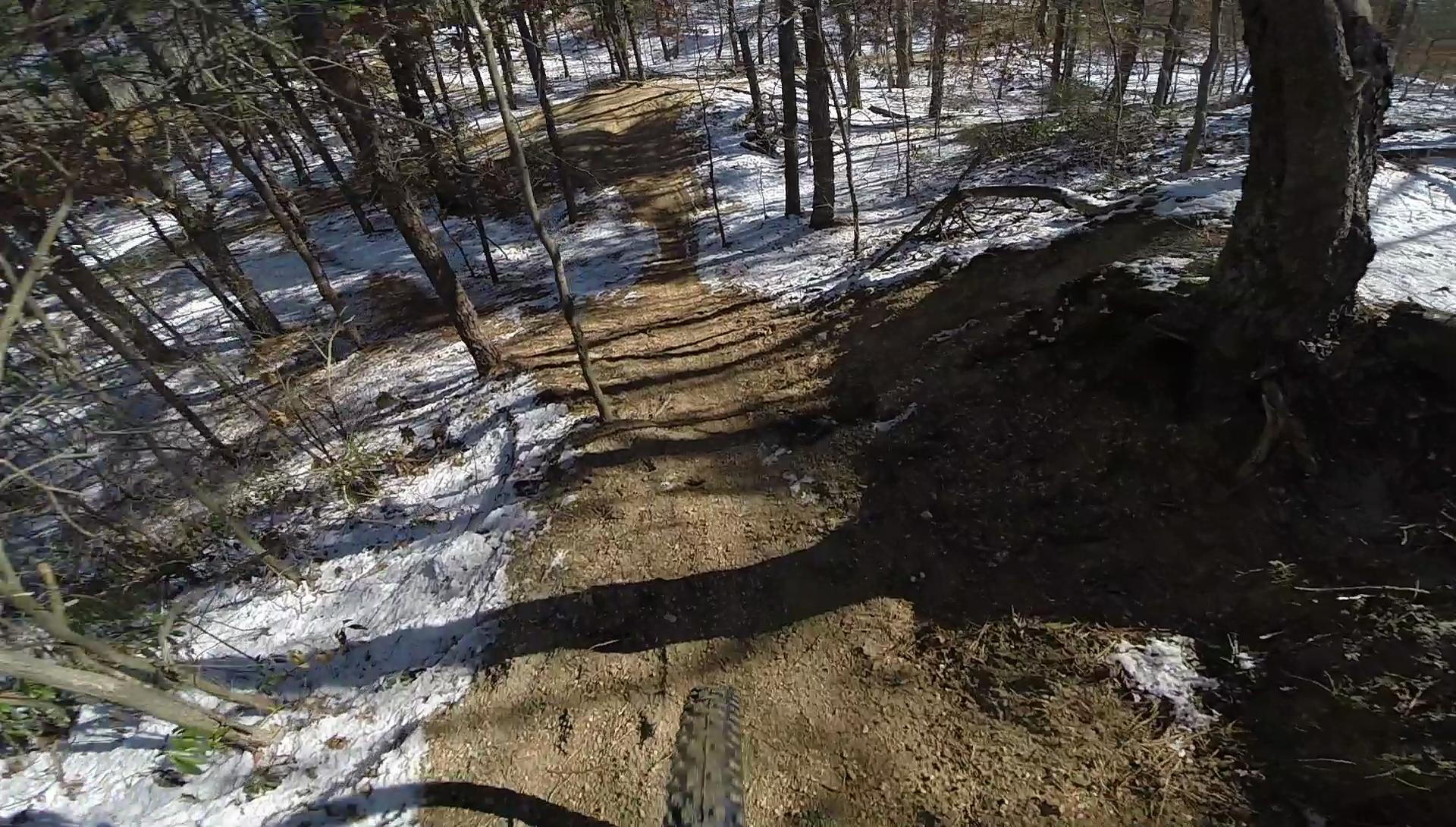 A mountain biking trail winding through a snowy forest, featuring a dirt path lined with trees and shadows cast on the ground. The perspective shows a view from above, highlighting the slope of the trail and the surrounding nature. Allaire State Park mountain bike trail.