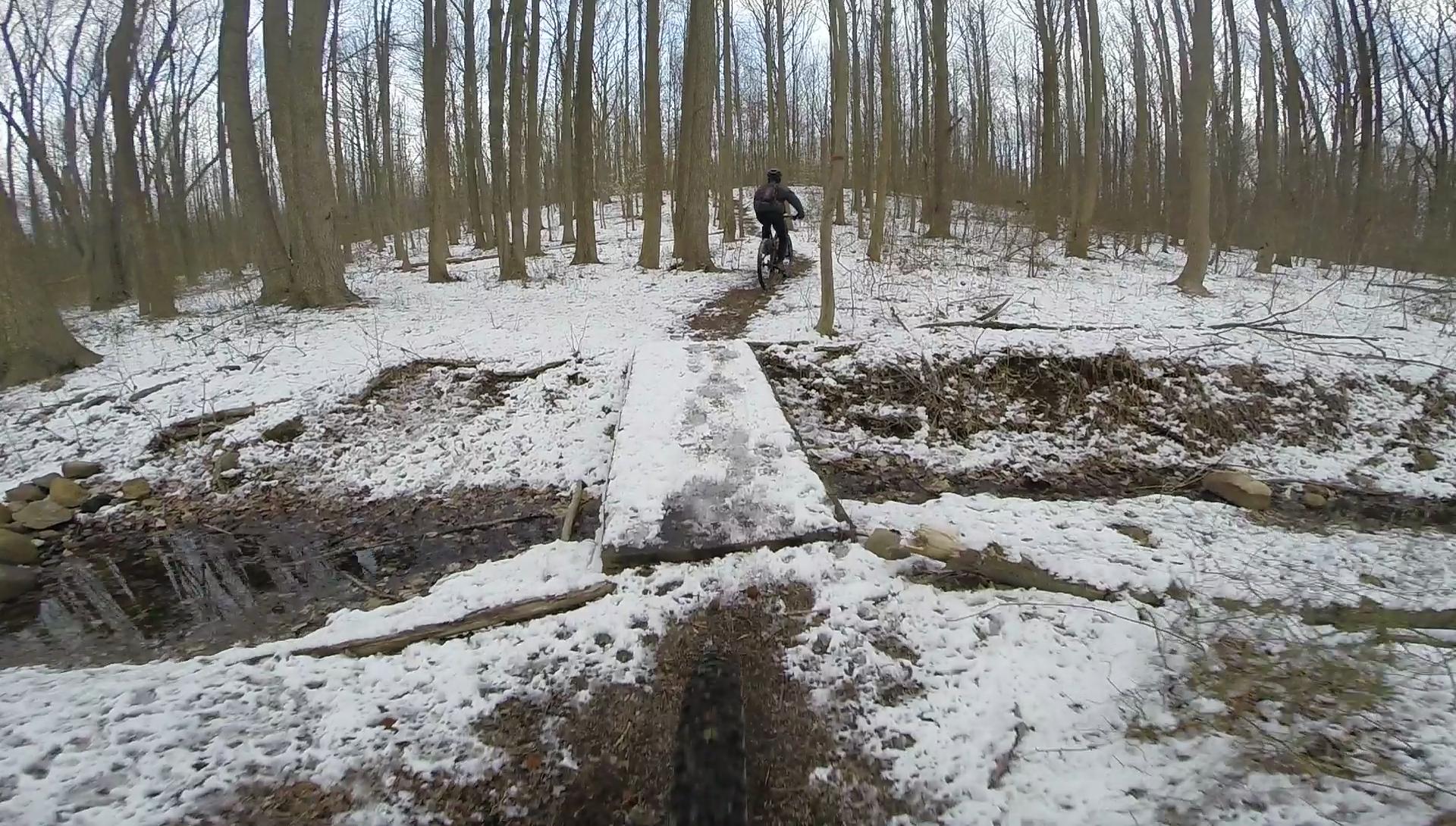 A mountain biker navigating a snowy forest trail over a wooden bridge, with trees in the background and a stream visible beneath the bridge. Wolfes Pond park mountain bike trail.