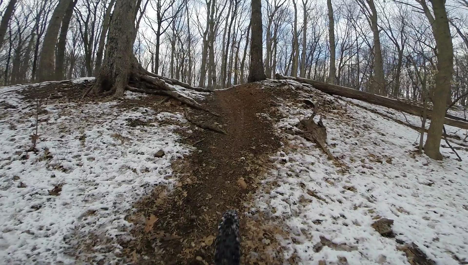 A view of a muddy, snow-covered trail winding up a hillside in a forest, with bare trees and a cloudy sky visible in the background. The perspective captures the ground and a bicycle tire, emphasizing the off-road cycling environment. Wolfes Pond park mountain bike trail.