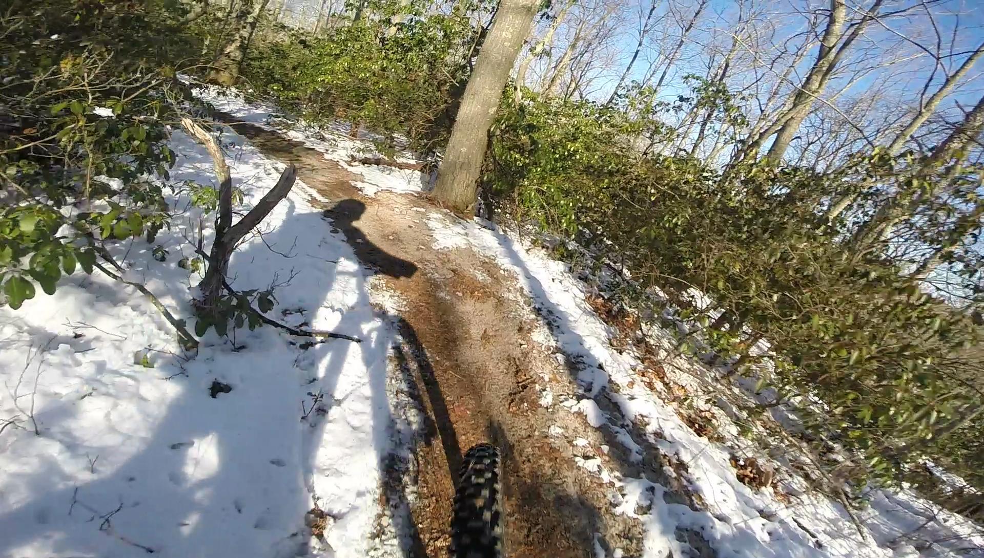 A mountain biker riding on a snowy trail surrounded by trees and greenery, with the bike's shadow visible on the path. The scene captures the contrast between the snow and the earthy trail under a clear blue sky. Allaire State Park mountain bike trail.