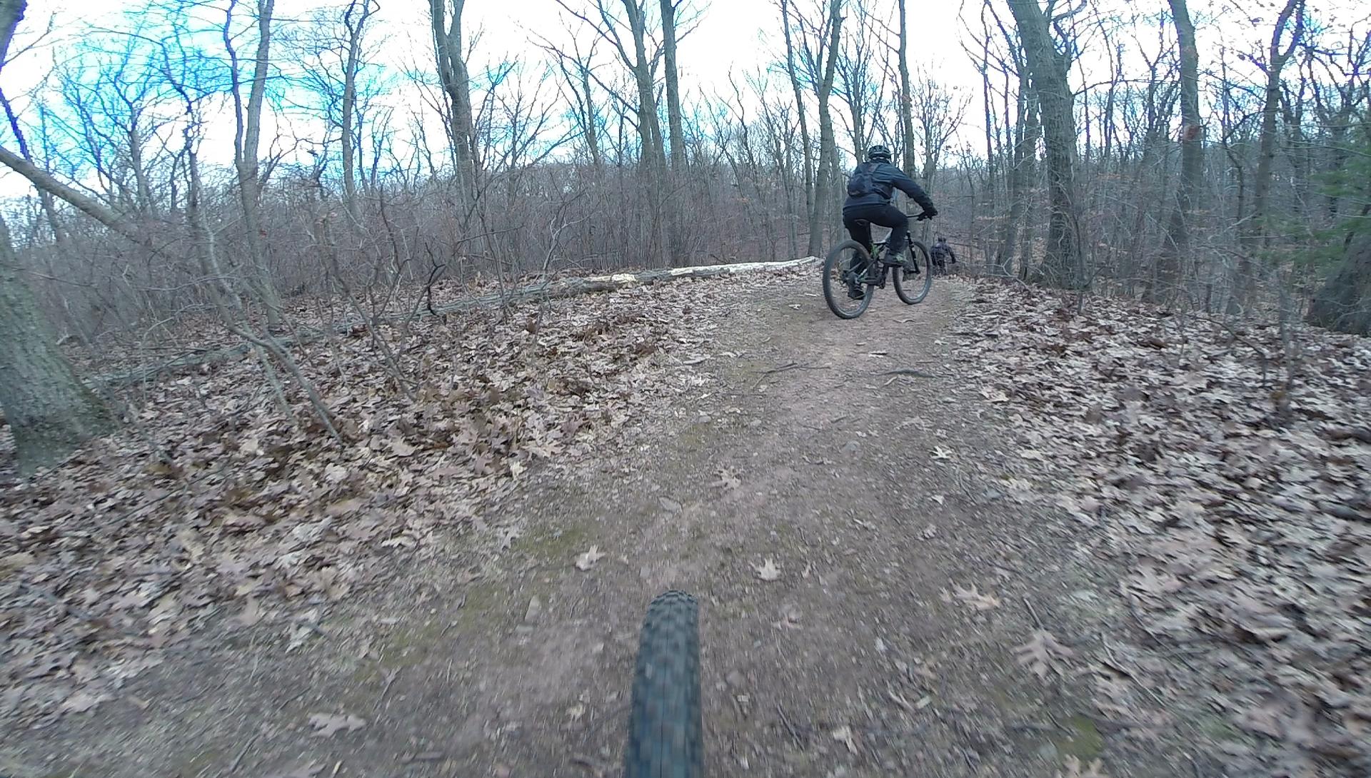 A view of a dirt biking trail in a wooded area. The image shows two cyclists riding on a path covered with fallen leaves, surrounded by bare trees in a winter setting. One cyclist is in the foreground with their bike tire in view, while the other cyclist is in motion further along the trail. The sky is partly cloudy. Richmond Avenue and Forest Hill road mountain bike trail.