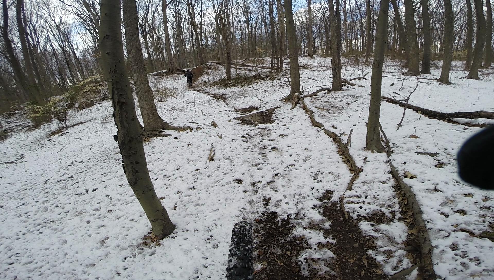 A snowy forest trail with scattered snow covering the ground and remnants of autumn leaves. In the background, a person is walking along the path, surrounded by bare trees. The trail is slightly muddy, with visible tire tracks and footpaths leading through the snow. The sky is partly cloudy, creating a wintery atmosphere. Wolfes Pond park mountain bike trail.