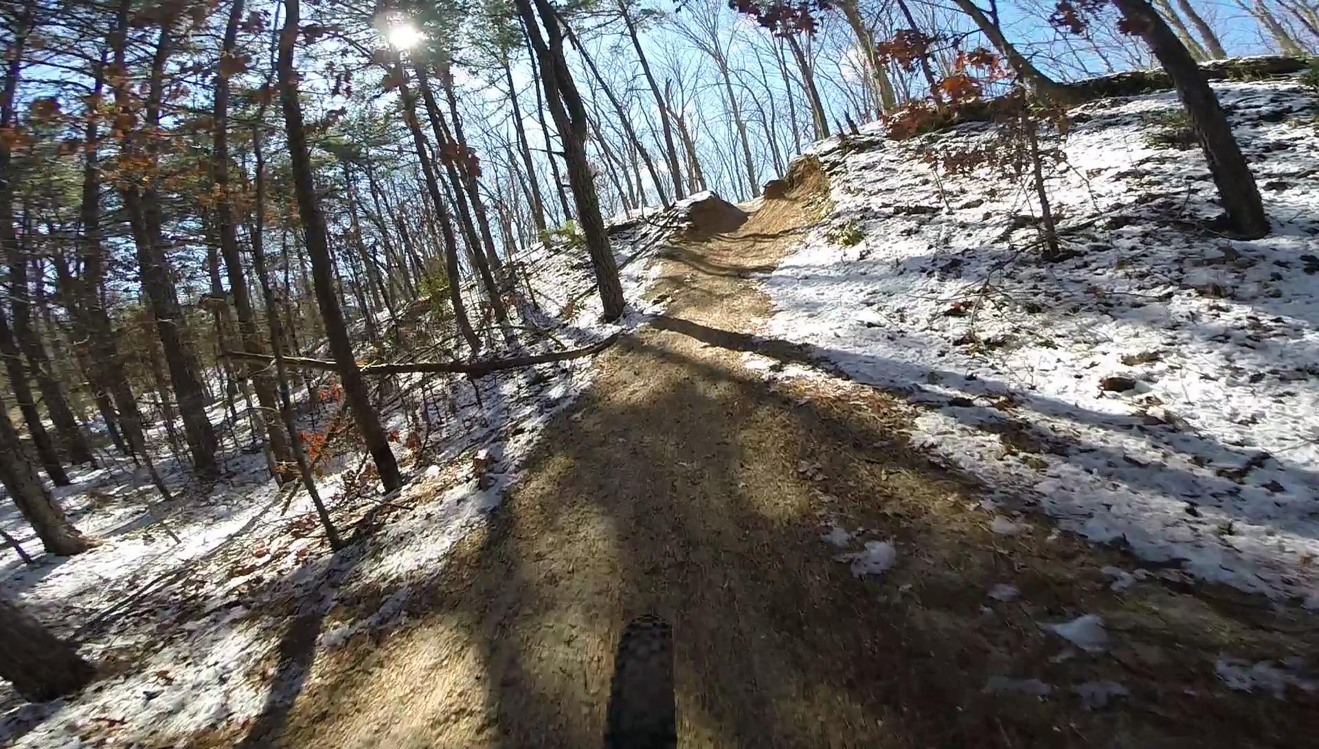 A dirt trail winding through a forest, with sunlight filtering through trees and patches of snow along the path. The trail has an incline with shadows casting from the trees, creating a serene winter landscape. Allaire State Park mountain bike trail.