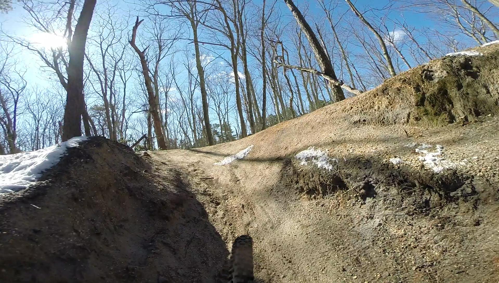 A dirt bike trail winding through a forest, with bare trees under a clear blue sky. Snow patches are visible along the trail, indicating a cooler season. The perspective shows the trail from a low angle, highlighting the texture of the dirt path. Allaire State Park mountain bike trail.