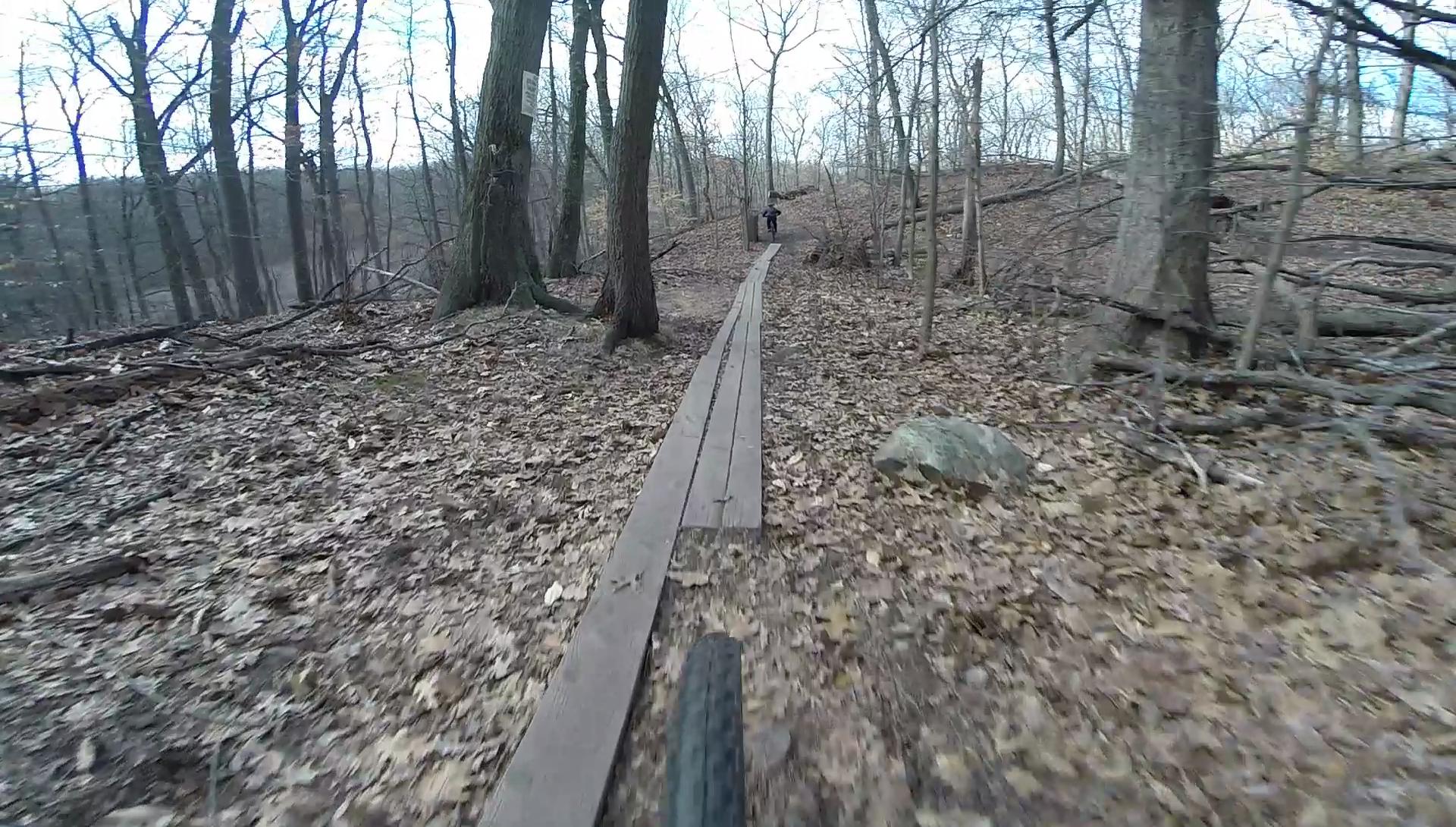 A mountain biking trail winding through a wooded area during early spring, featuring a wooden plank pathway over a carpet of fallen leaves. A cyclist is seen in the distance, navigating the trail among bare trees and scattered stones. Richmond Avenue and Forest Hill road mountain bike trail.
